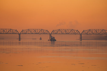 Tugboat passing under railway bridge at sunriseの写真素材