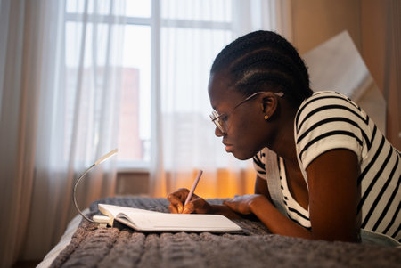 Young black woman lying on bed writing in notebook by lamp lightの写真素材