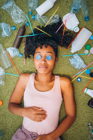 Young woman lying on green carpet covered with plastic trash and bottle caps on eyes