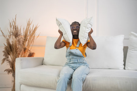 Young woman covering ears with pillows on sofa in living roomの写真素材