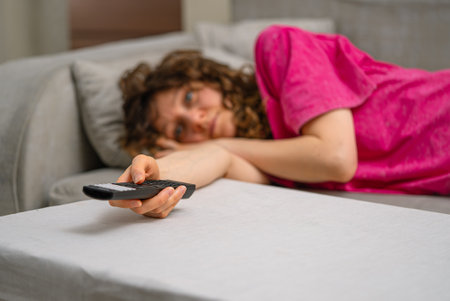 Frustrated female reclining on gray couch, mindlessly changing tv channels, seeking entertainment during laid-back home leisure timeの写真素材
