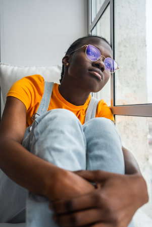 Black woman with glasses sitting near window, looking outside with pensive expression, embodying quiet reflection and urban solitudeの写真素材
