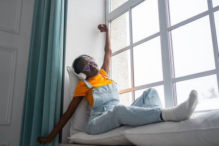 African American woman sitting near expansive window, wearing headphones and sunglasses, stretching with tranquil contentment in soft morning lightの写真素材