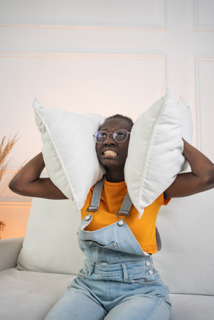 Black woman blocking ears with pillows while sitting on couch, displaying frustration from overwhelming noise and seeking quiet moment of solitudeの写真素材