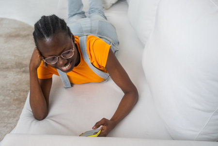 Smiling woman with braided hair and glasses relaxing on white sofa, browsing smartphone in comfortable casual outfitの写真素材