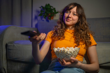 Young woman with curly hair relaxing on sofa, holding remote and popcorn bowl, watching late-night television with comfortable expressionの写真素材