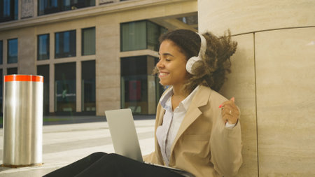 A young woman enjoys a moment of relaxation, smiling as she listens to music on her headphones and uses her laptopの写真素材