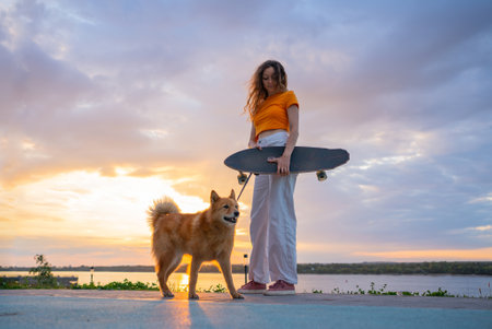 Young woman holding skateboard posing with dog by river at sunsetの写真素材