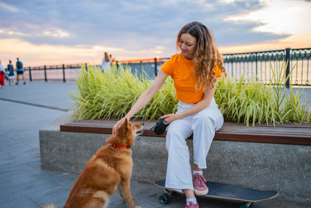 Young woman petting her dog while sitting on her skateboard by the river at sunsetの写真素材