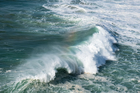 Massive ocean wave crashing near rocky coastline, generating dramatic water spray with colorful rainbow arcing above turbulent seascapeの写真素材