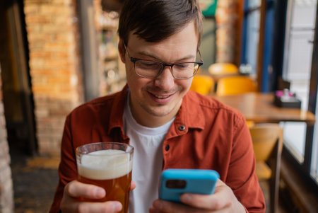 Bearded man relaxing at pub bar, drinking cold beer and scrolling smartphone, radiating casual contentment during leisurely momentの写真素材