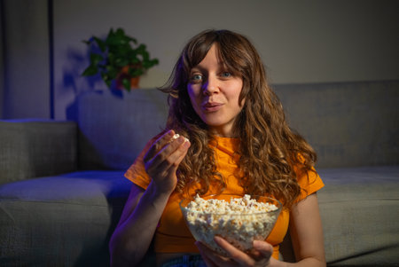 Woman sitting cross-legged on floor, eating popcorn near couch, watching movie with relaxed expression during casual home entertainment eveningの写真素材
