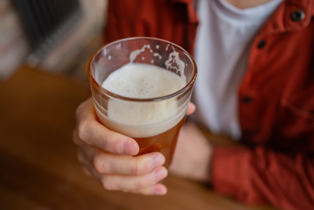 Glass of craft beer with foamy head, held by hands in warm pub setting, showcasing relaxed enjoyment and beverage craftsmanshipの写真素材