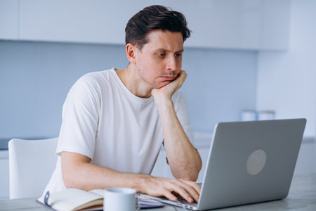 Home office professional leaning on hand, looking fatigued while typing on laptop during remote work session, expressing workplace wearinessの写真素材