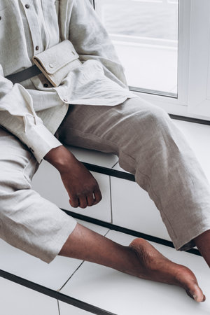 Trendy person leaning casually on white stairs, wearing neutral linen outfit accented by beige belt bag, embodying understated urban chic styleの写真素材