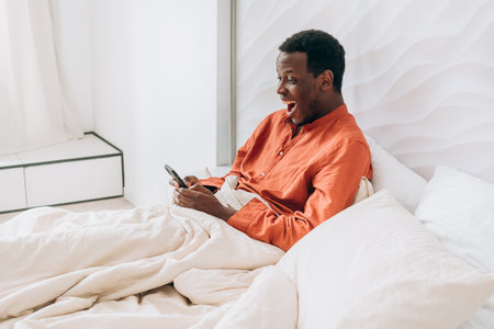 Surprised male resting under soft blanket, browsing smartphone with wide-eyed expression near sunlit bedroom window, capturing morning relaxationの写真素材