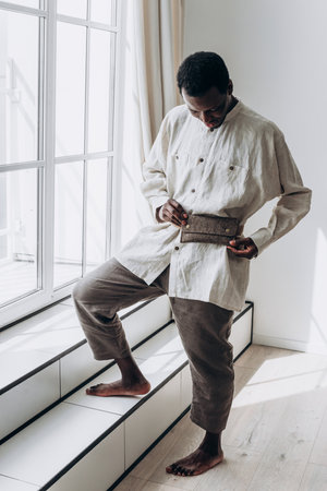 Barefoot african american male model in linen attire and fanny pack, leaning near window, basking in soft natural sunlight filtering through contemporary apartment interiorの写真素材