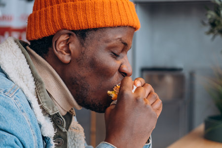 Young black man with orange winter hat enjoying eating a burger in a fast food restaurant, side view close-up, concept of junk food, street food, and winter seasonの写真素材