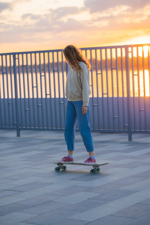 Young woman riding longboard beside river during golden sunset, embodying carefree movement and urban lifestyle with serene landscape backgroundの写真素材