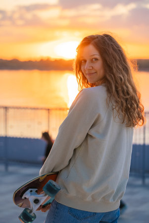 Young woman with curly hair gripping skateboard, turning head and smiling at sunset near river, golden light illuminating urban landscape with youthful energyの写真素材
