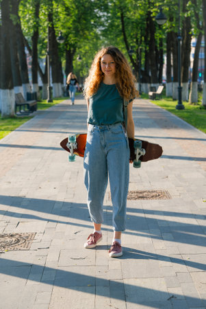Curly-haired woman carrying longboard, walking through sunlit urban park, embodying carefree street style and outdoor freedomの写真素材