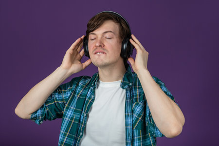 Young man in plaid shirt listening to music, adjusting headphones with closed eyes, standing against vivid purple backgroundの写真素材