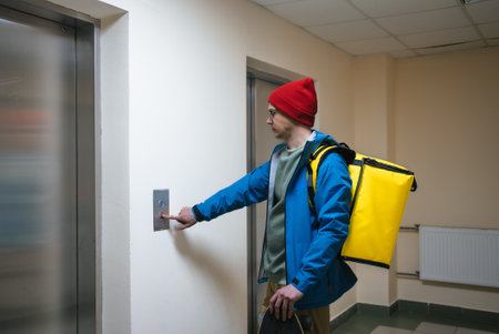 Food delivery worker pressing elevator button, wearing casual attire and carrying yellow thermal backpack in office building hallway, facilitating quick meal transportの写真素材