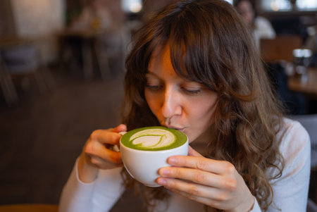 Young woman enjoying matcha latte, gently blowing on vibrant green surface with intricate latte art in warm, inviting cafe settingの写真素材