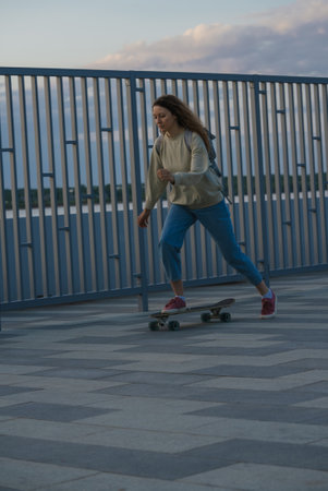 Young woman longboarding along riverside, enjoying evening cityscape, flowing with urban energy and sense of freedomの写真素材