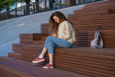 Curly-haired woman sitting on wooden steps, smartphone in hand, skateboard nearby, navigating digital world within urban park settingの写真素材
