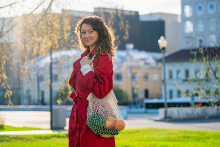Woman wearing red coat, carrying mesh bag with fresh produce, walking through urban park, exemplifying sustainable lifestyleの写真素材