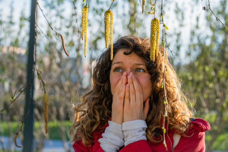 Curly-haired woman in red coat blocking nose and mouth while experiencing seasonal allergy symptoms near blooming birch trees during spring season in urban parkの写真素材