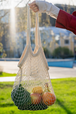 Woman holding reusable mesh bag with fresh groceries, standing in urban park, representing sustainable shopping and eco-friendly lifestyle choicesの写真素材