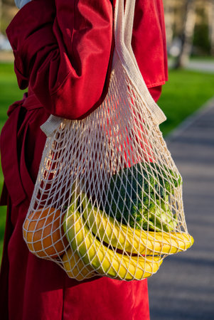 Red-coated woman carrying reusable string bag with fresh produce, highlighting eco-friendly grocery shopping and sustainable lifestyle choicesの写真素材