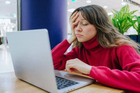 Young woman in red turtleneck feeling tired and stressed while working on her laptop in a coworking space, experiencing burnout and frustration from a challenging design orderの写真素材