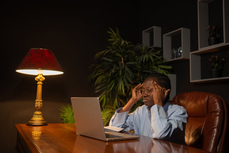 Stressed african american professional experiencing workplace burnout, pressing fingertips against temples while managing overwhelming work pressureの写真素材