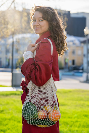 Young woman with windblown curly hair wearing bright red coat carrying mesh grocery bag filled with colorful fresh produce while walking urban streetの写真素材