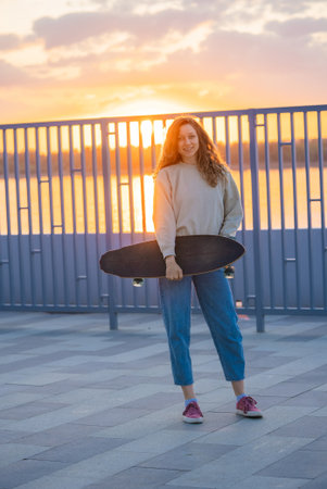 Curly-haired female resting on longboard during golden sunset, silhouetted against metal fence, capturing urban skateboarding freedom and relaxed evening moodの写真素材