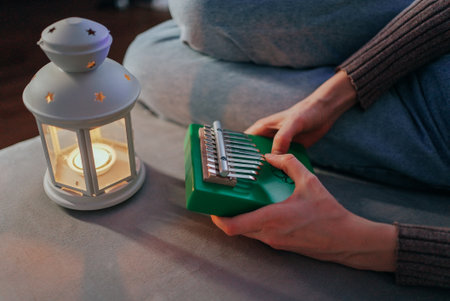 Female musician playing green kalimba near candle lantern, creating intimate acoustic moment with soft, meditative musical instrument on cozy sofaの写真素材