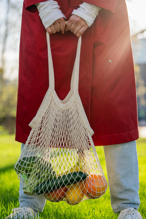 Woman carrying eco-friendly mesh bag overflowing with organic produce, representing sustainable shopping and environmental responsibilityの写真素材