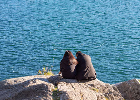 Backside view of the couple sits on rocks on sea shore close to each other in the background of the beautiful blue sea. Selective focus, travel photo, street view.の写真素材