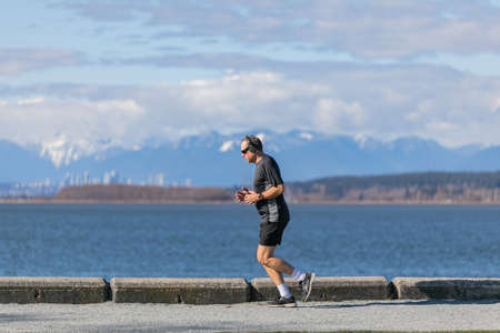 Old active man running in the park wearing sunglasses and listening music in his headphones in the beautiful nature background. White Rock, BC, Canada-March 15,2021. Street view, travel photoのeditorial素材