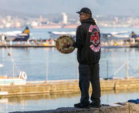 Rear view of native american man with sacred native frame drum worshiping by the shore in the Harbor of Vancouver, BC, Canada-November 1,2020. Travel photo, street view, selective focus.のeditorial素材