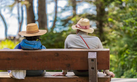 Rear view of a senior couple wearing hats sitting on a wooden bench in the park. Vancouver Island, BC, Canada-August 9,2021. Street view, travel photo, selective focus, blurred.の写真素材