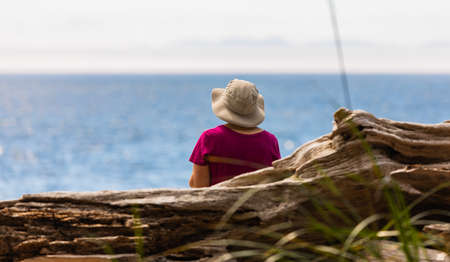 Rear view of alone senior woman with hat sitting on the beach looking at the blue water of the sea. Vancouver Island, BC. Street view, travep photo, selective focus, blurredの写真素材
