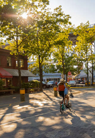 Young woman tourist with backpack and hat enjoys riding her small bicycle in the city at sunset. street view, travel photo, selective focus, blurredの写真素材