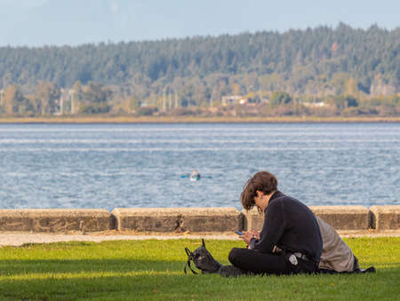 Two freinds with a smartphone in their hands are sitting on the grass. Young people busy with their phones relaxing in a parkのeditorial素材