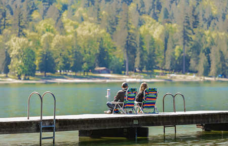 Couple sitting on dock at the beautiful lake. Rear view of a romantic young couple sitting on pier enjoying stunning views at the Cultus Lake BC, Canada-May 22,2022. Travel photography, selective focusのeditorial素材