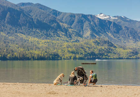 Family of three people sitting on the shore of a lake in summer and watching the beautiful view with dog. Back view, travel photo, selective focus-May 22,2022-Cultus Lake BC Canadaのeditorial素材