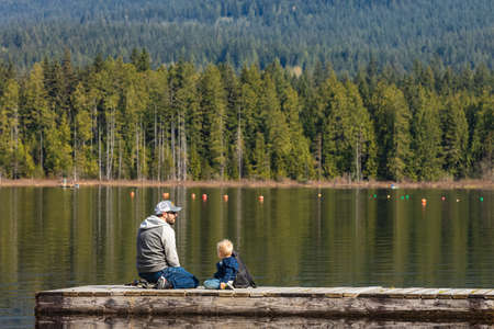 Father with son sitting on the wooden pier on a mountain lake. Travel photo, selective focus, copy space for text-April 24,2022-Whonnock Lake BC, Canadaのeditorial素材